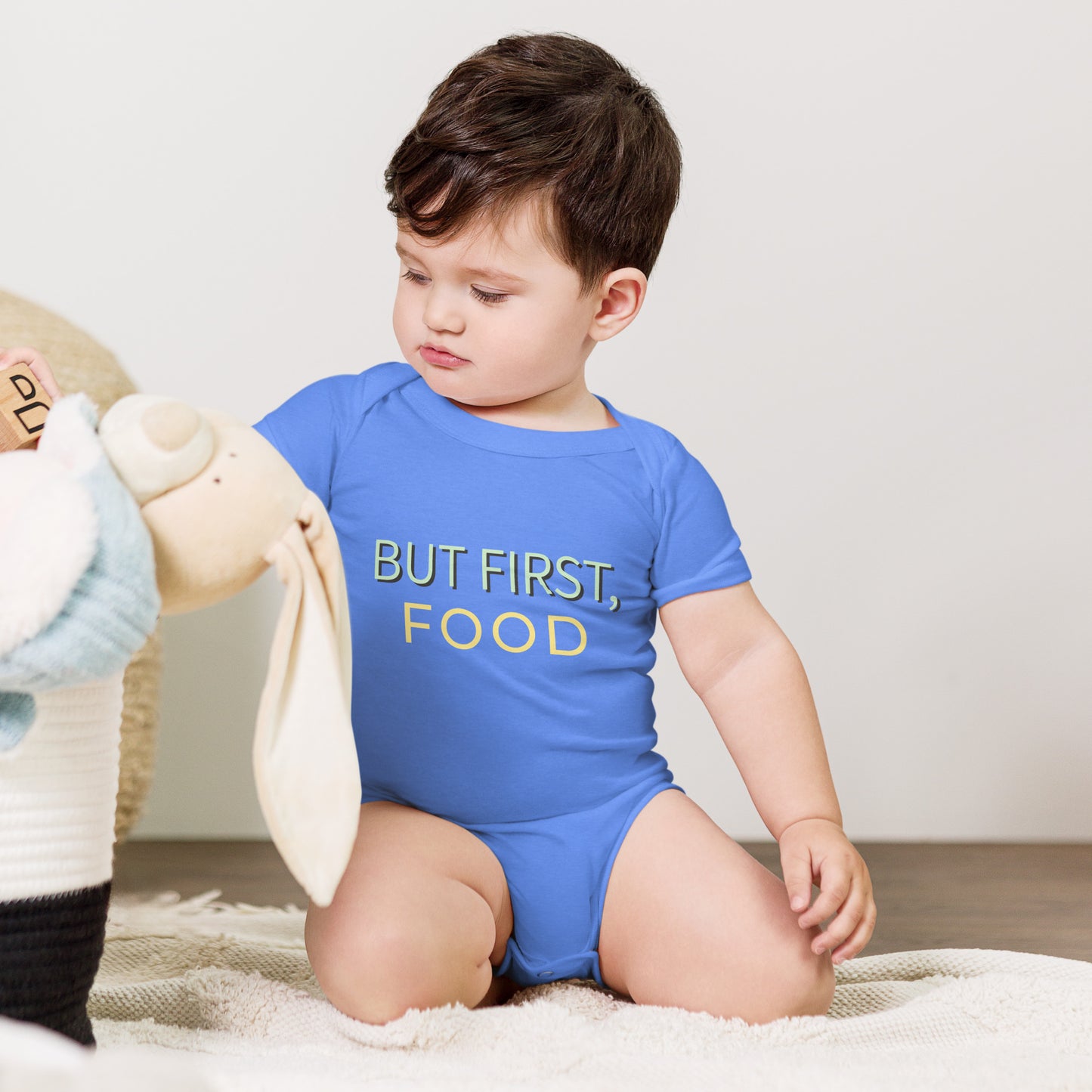 Experience the cuteness overload of a baby dressed in a blue bodysuit proudly declaring, "But first food." Cherish the joy of early feeding milestones.