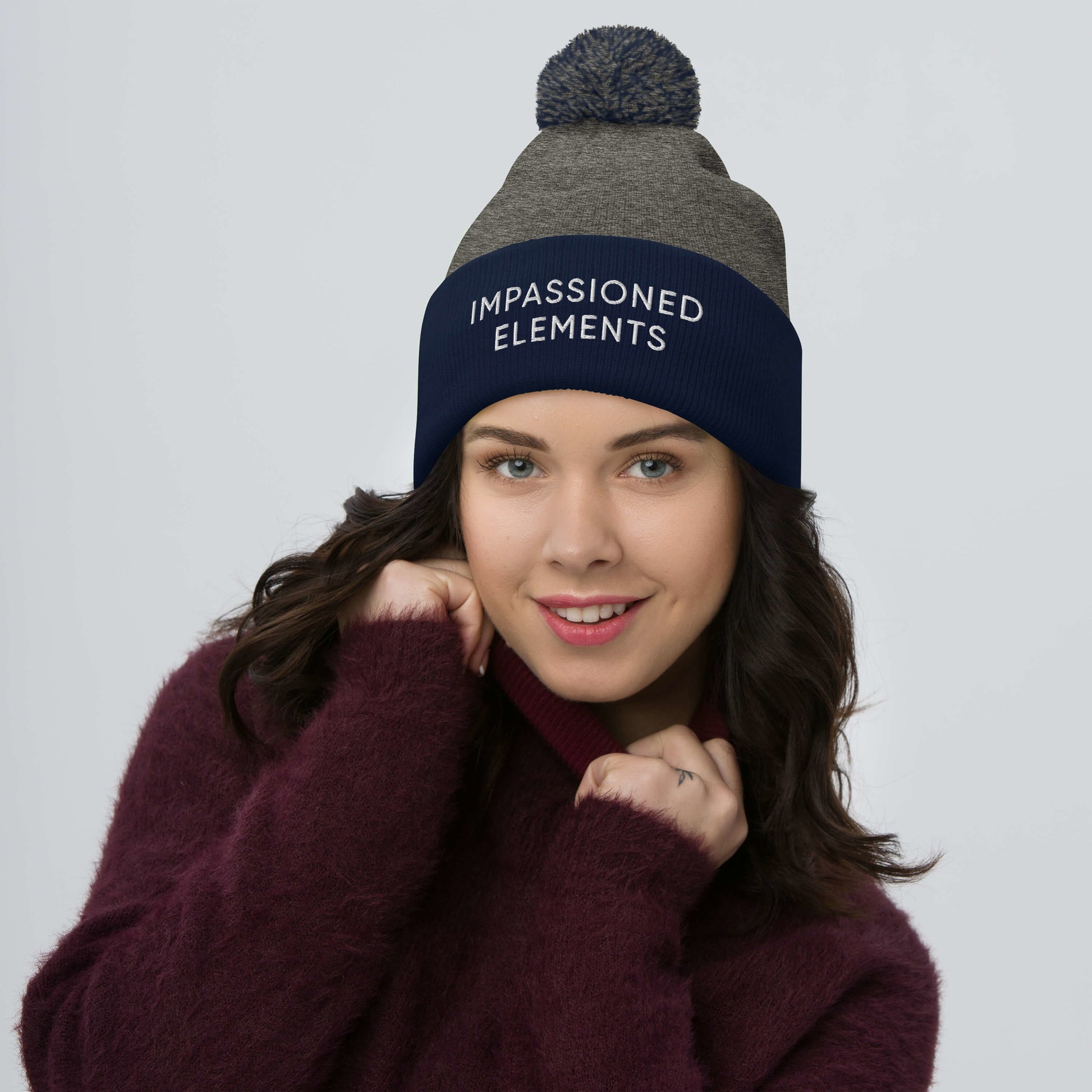 Bold woman wearing a dark heather grey and navy pom-pom embroidered Impassioned Elements beanie with flair